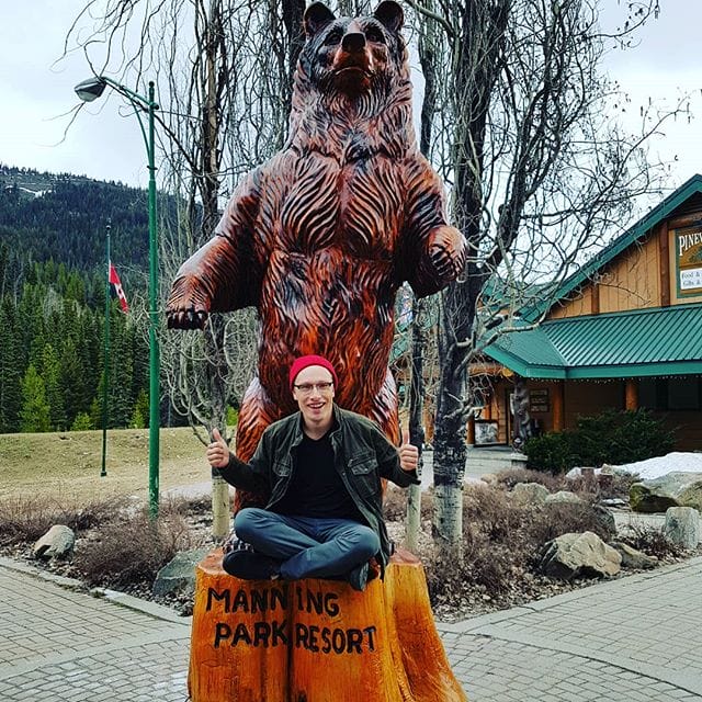 A person sits on a wooden sculpture labeled "Manning Park Resort" with a large bear statue behind them; trees, campsites, a building, and mountains are in the background.