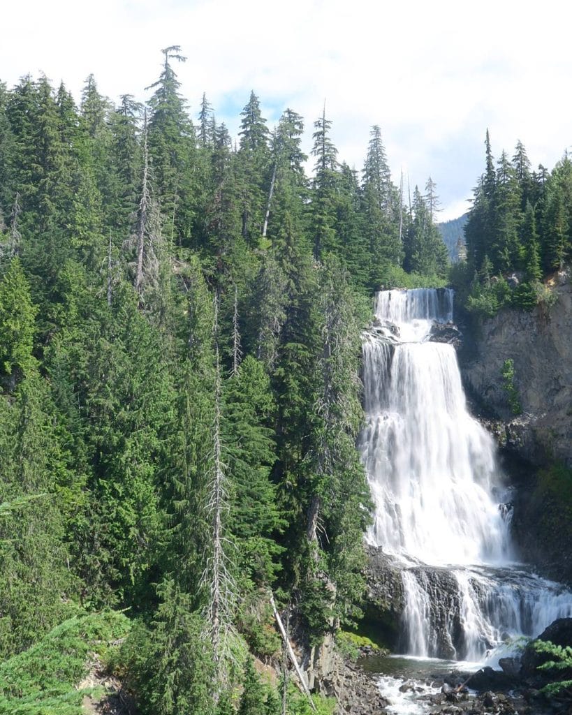 A multi-tiered waterfall flows through a dense forest of evergreen trees under a partly cloudy sky, capturing the serene beauty of Whistler waterfalls.