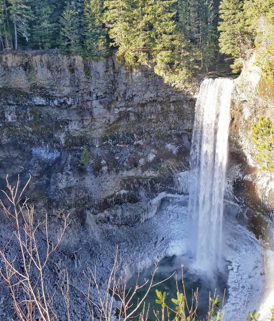 A tall waterfall flows over a rocky cliff surrounded by evergreen trees, with a pool and some ice at the base—one of the stunning Whistler waterfalls.