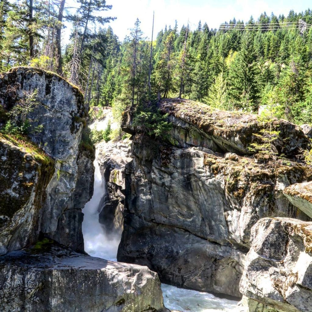 A waterfall flows between large rocky cliffs surrounded by dense evergreen forest under a bright, sunny sky, capturing the natural beauty of Whistler waterfalls.