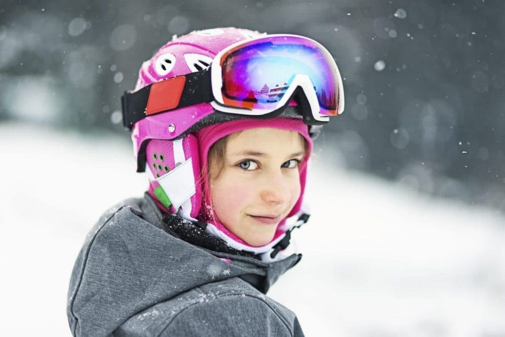 A child wearing a pink ski helmet and goggles looks at the camera, standing outdoors in a snowy environment.