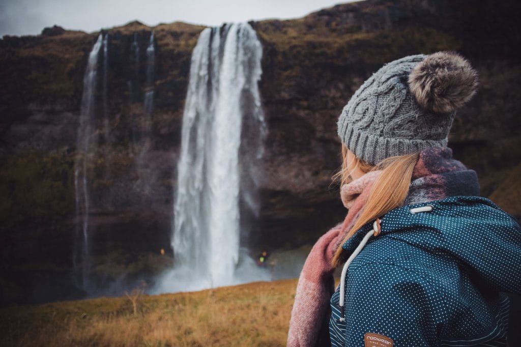 A person in a winter coat and knit hat stands in front of the Whistler waterfalls, gazing at the majestic, cascading water.