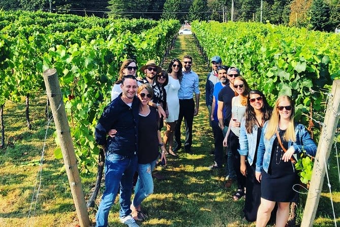 A group of people poses and smiles for a photo while standing in a single line between rows of grapevines at one of the scenic Fraser Valley Wineries on a sunny day.