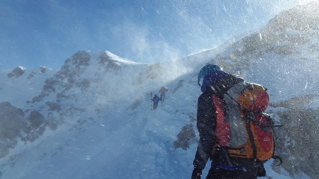 A group of climbers wearing winter gear ascend a snow-covered mountain ridge in windy, snowy conditions.
