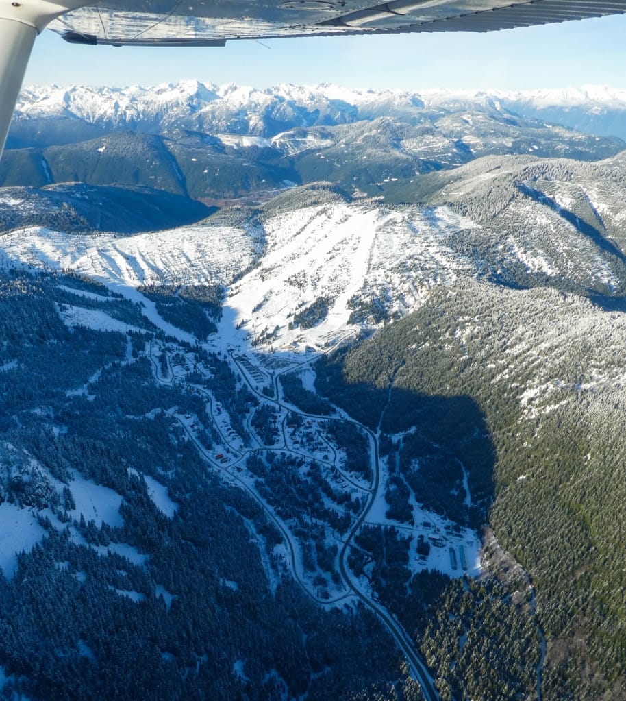 Aerial view of a snow-covered mountain landscape with winding roads, forested areas, and scattered buildings near ski resorts in the Fraser Valley, taken from an aircraft.