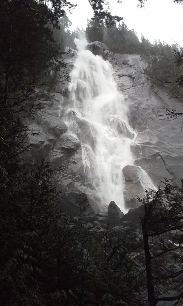 A tall waterfall cascades down a rocky cliff surrounded by dense trees under an overcast sky, reminiscent of the serene beauty found at Whistler waterfalls.