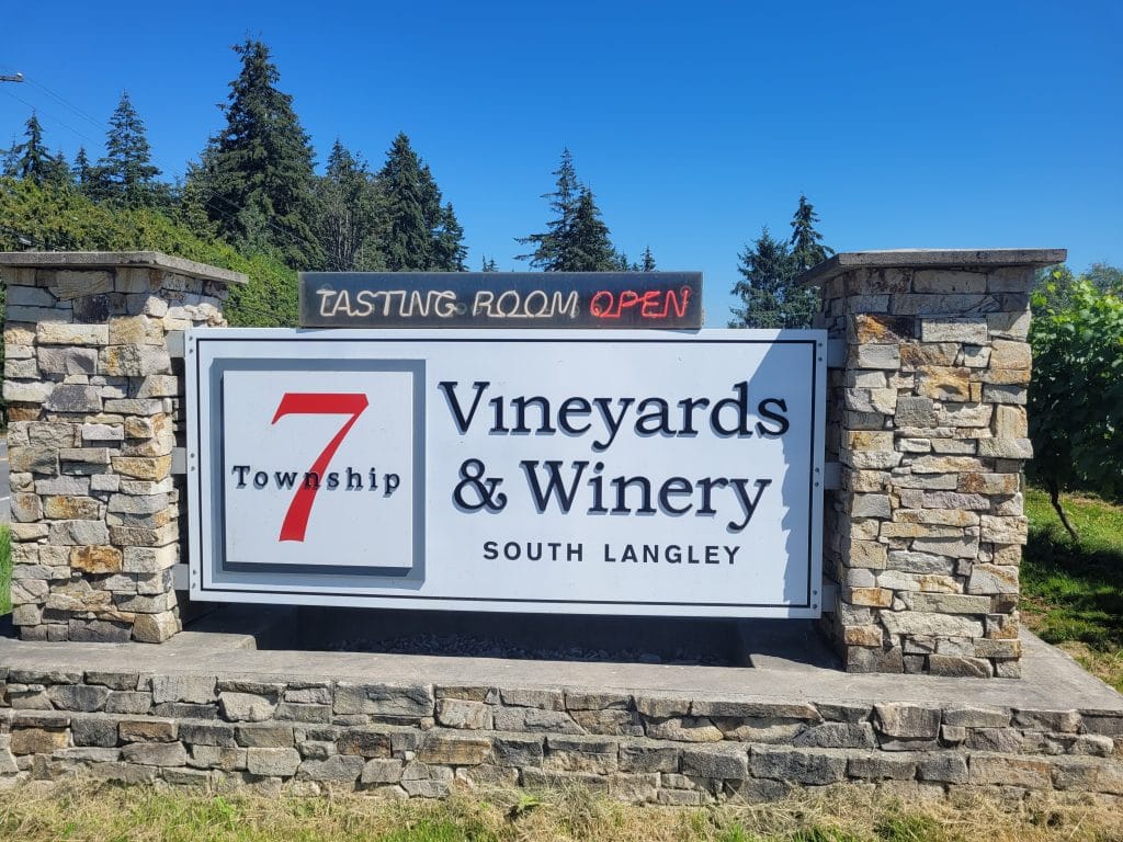 A stone sign reads "Township 7 Vineyards & Winery South Langley" with a smaller sign above stating "TASTING ROOM OPEN." Nestled among trees and blue sky, this spot is a standout among Fraser Valley Wineries.