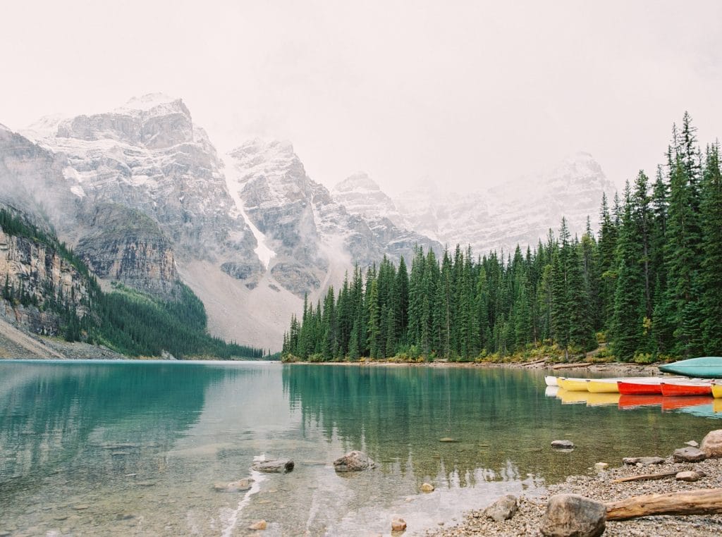 A clear turquoise lake surrounded by pine trees and snow-capped mountains, with colorful canoes on the shore in the foreground.