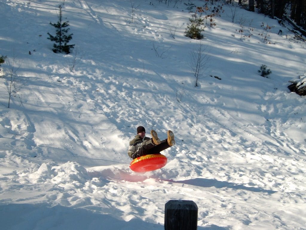 A person rides a bright orange snow tube down a snowy hill at Sasquatch Mountain Resort, feet lifted in the air, surrounded by trees and snow-covered ground.