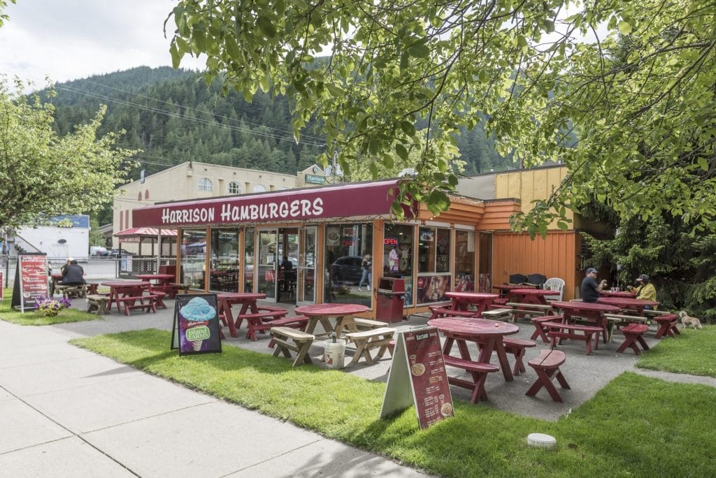 Outdoor view of Harrison Hamburgers, known as one of the best restaurants in Harrison, with red picnic tables on a grassy area under leafy trees, set against a backdrop of mountains on a cloudy day.