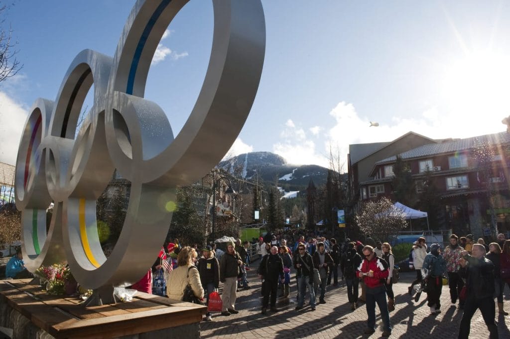 A crowd gathers near large Olympic rings in an outdoor plaza in Whistler, with mountains and buildings in the background on a sunny day—a popular spot for activities Whistler is known for.