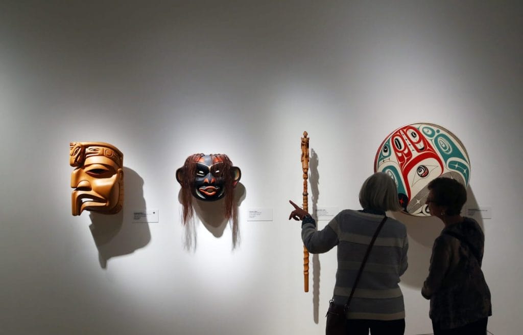 Two people explore activities Whistler offers as they view Indigenous masks, a staff, and a painted round plaque displayed on a white gallery wall under focused lighting.