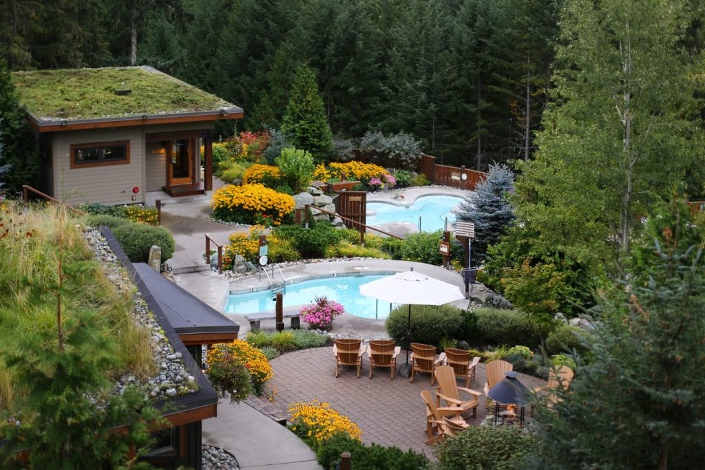 A landscaped outdoor spa in Whistler features hot tubs, lounge chairs, and garden paths surrounded by lush trees and colorful flowers—perfect for relaxing after your activities Whistler has to offer.