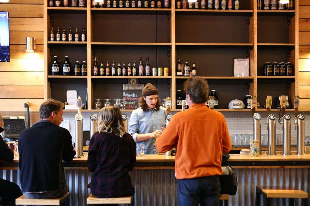 A bartender serves drinks to three customers seated at a bar, with wooden shelves displaying bottles and glasses in the background—one of the cozy activities Whistler visitors can enjoy after a day on the slopes.