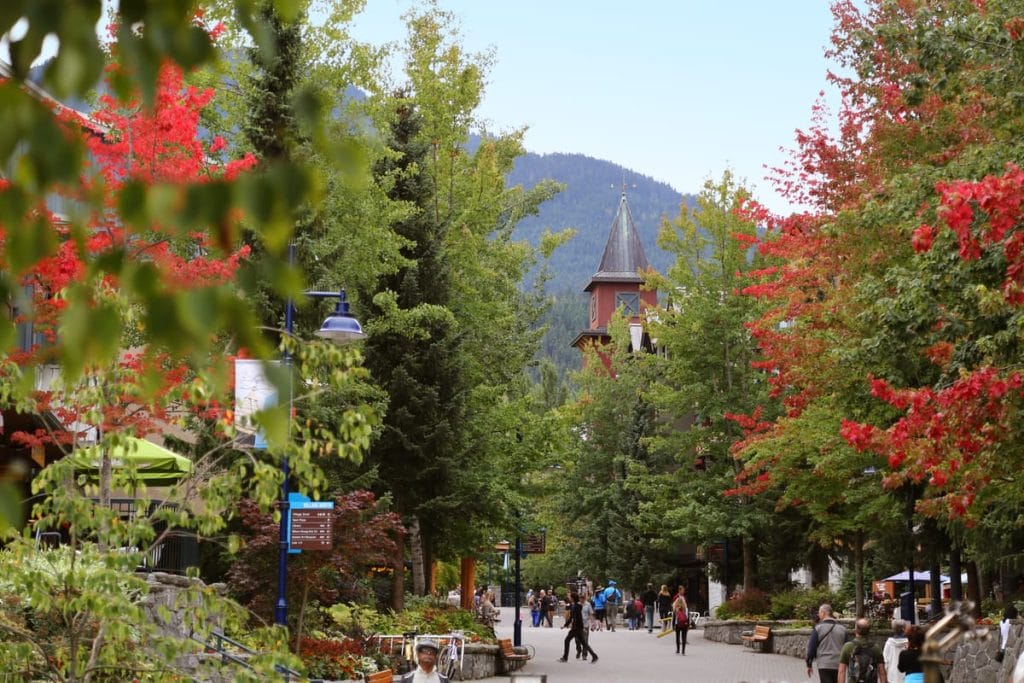Pedestrian pathway lined with green and red-leaved trees, people walking and enjoying activities Whistler is known for, with a church tower visible among the trees and mountains in the background.