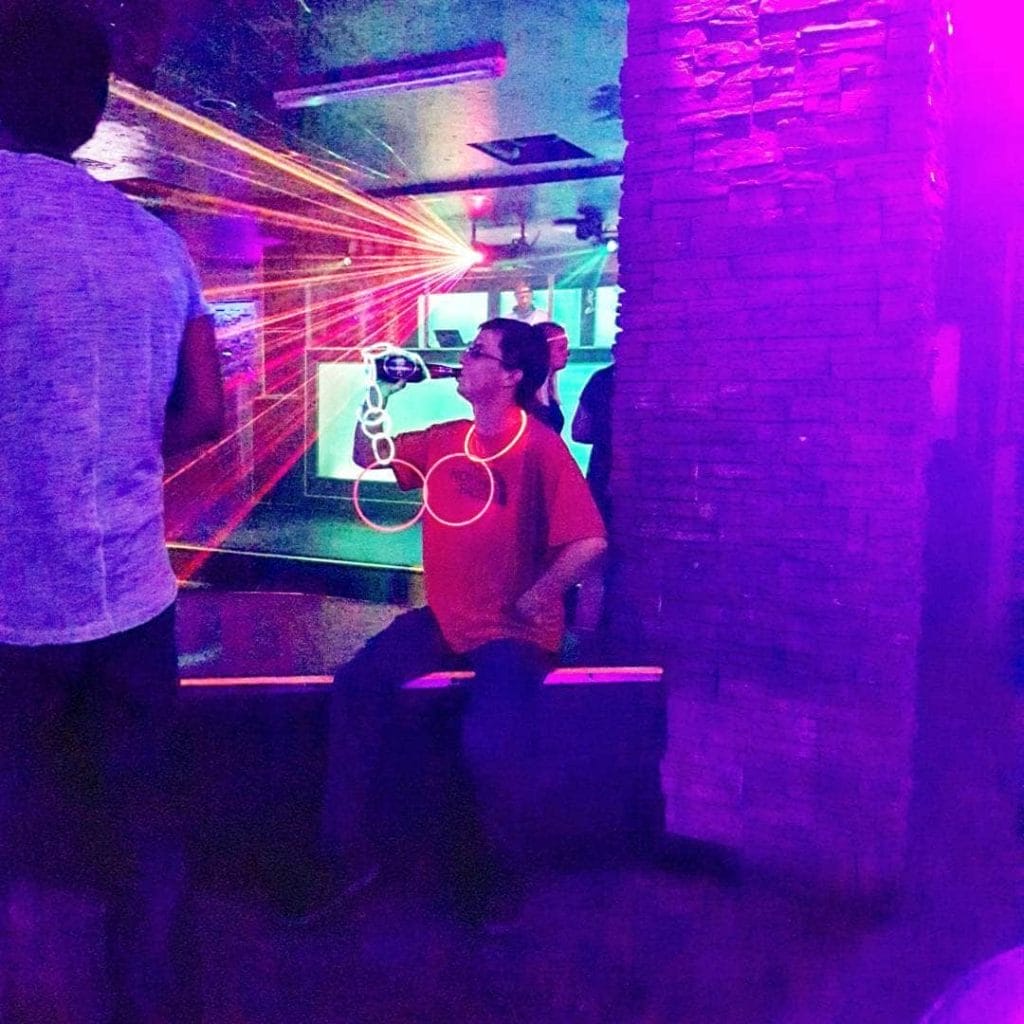 A man in a red shirt sits on a bench in a dimly lit room with purple lighting and laser lights, enjoying the vibrant Whistler nightlife as he drinks from a bottle.