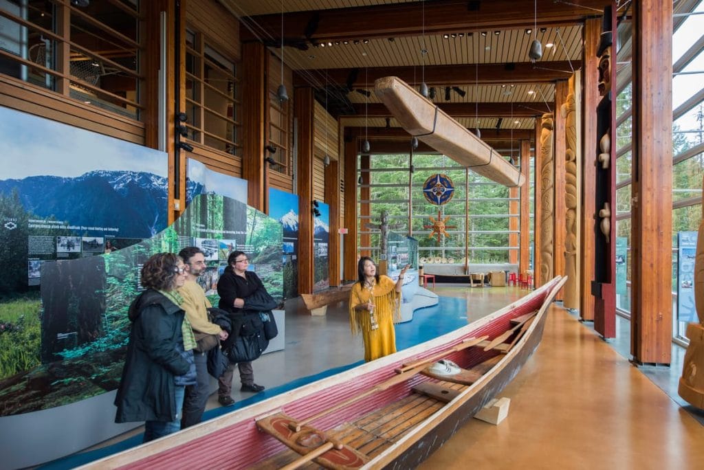 A museum guide speaks to a group in front of a traditional wooden canoe display, with large windows and forested scenery outside—a perfect glimpse into the cultural activities Whistler has to offer.