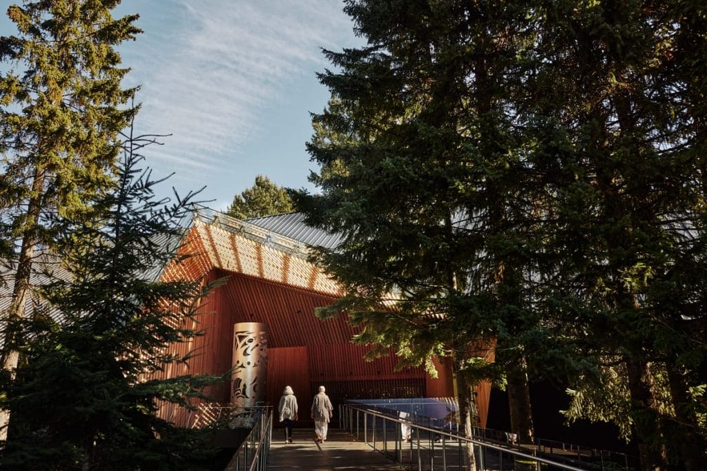 Two people walk on a ramp toward a wooden building surrounded by tall pine trees under a blue sky, enjoying outdoor activities Whistler is known for.