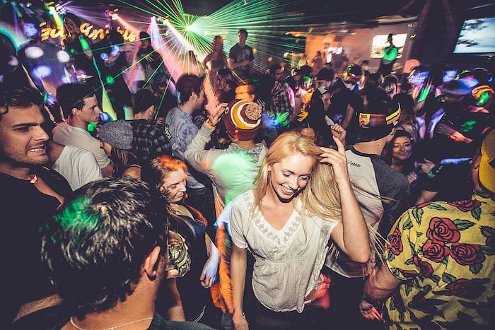 A crowded nightclub scene with people dancing under colorful lights and laser effects, capturing the energy of Whistler nightlife. A woman in a white shirt is smiling and dancing in the foreground.