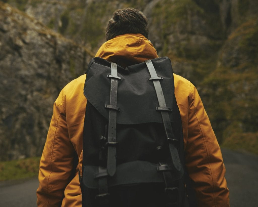 Person wearing a yellow jacket and black backpack stands on a road surrounded by rocky, green hills, facing away from the camera—ready to explore new adventures, just like discovering things to do in Cultus Lake.