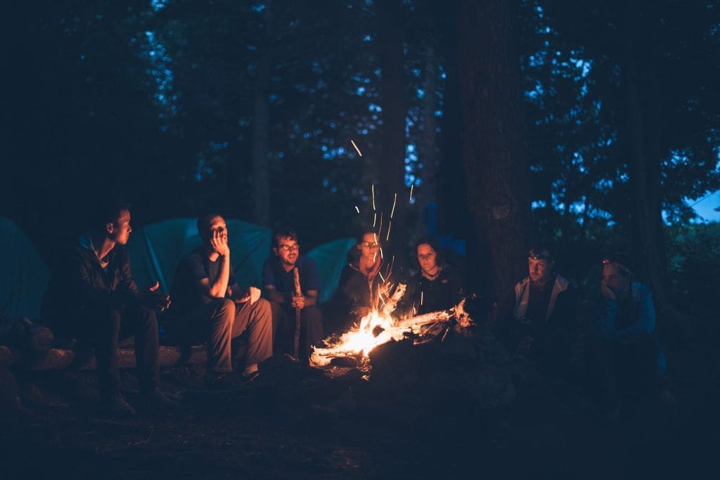 A group of people sit in a circle around a campfire at night in a wooded area with tents visible in the background, enjoying one of the classic things to do in Cultus Lake: camping under the stars with friends.