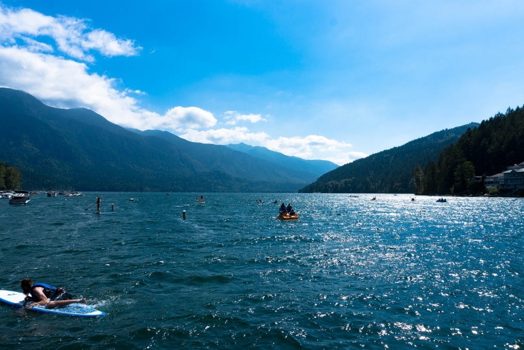 People paddle and kayak on a large lake surrounded by forested mountains under a blue sky with scattered clouds—perfect for those seeking fun things to do in Cultus Lake.