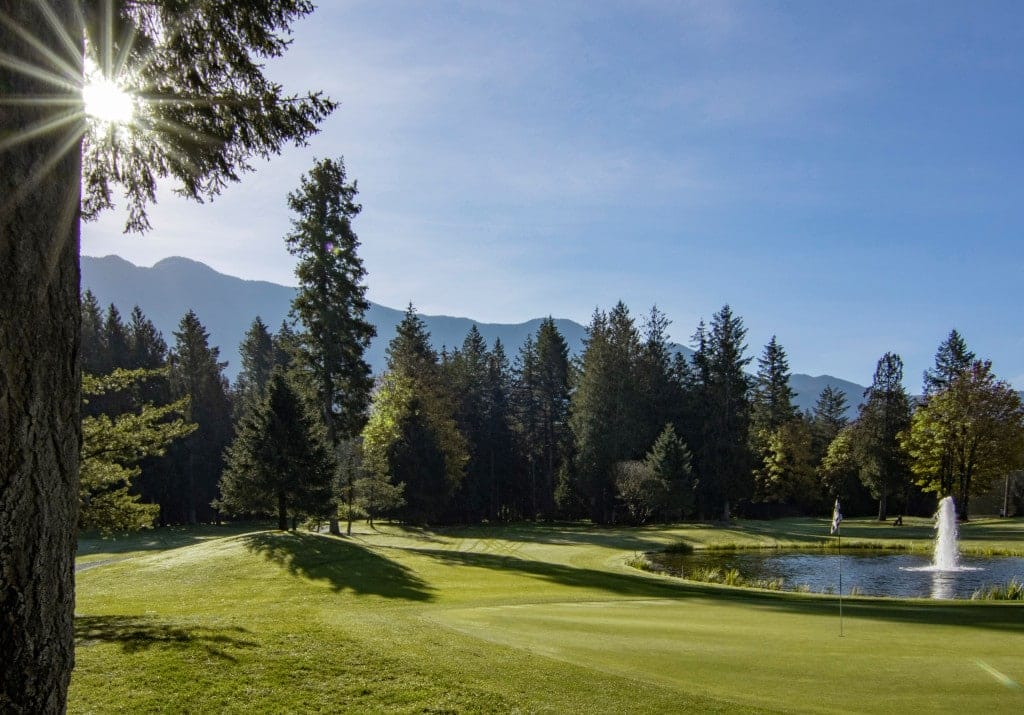 A beautiful golf course with a putting green, water fountain, trees, and mountains in the background, bathed in sunlight—one of the relaxing things to do in Cultus Lake.