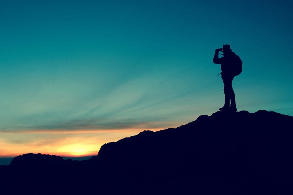 Silhouette of a person with a backpack, representing the adventurous Airbnb target market, standing on a rocky hilltop and looking through binoculars at sunset with a colorful sky in the background.