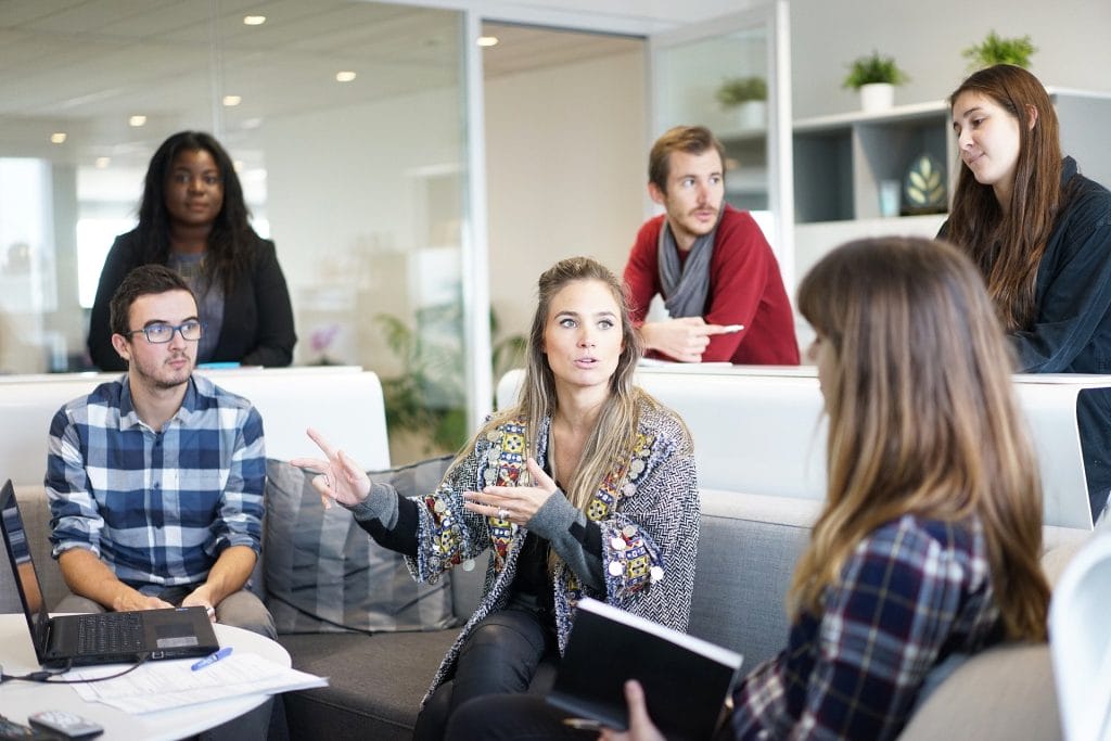 Six people in a modern office setting are engaged in a discussion about Airbnb market analysis. Some are sitting on couches while others are standing, with laptops and notebooks visible.