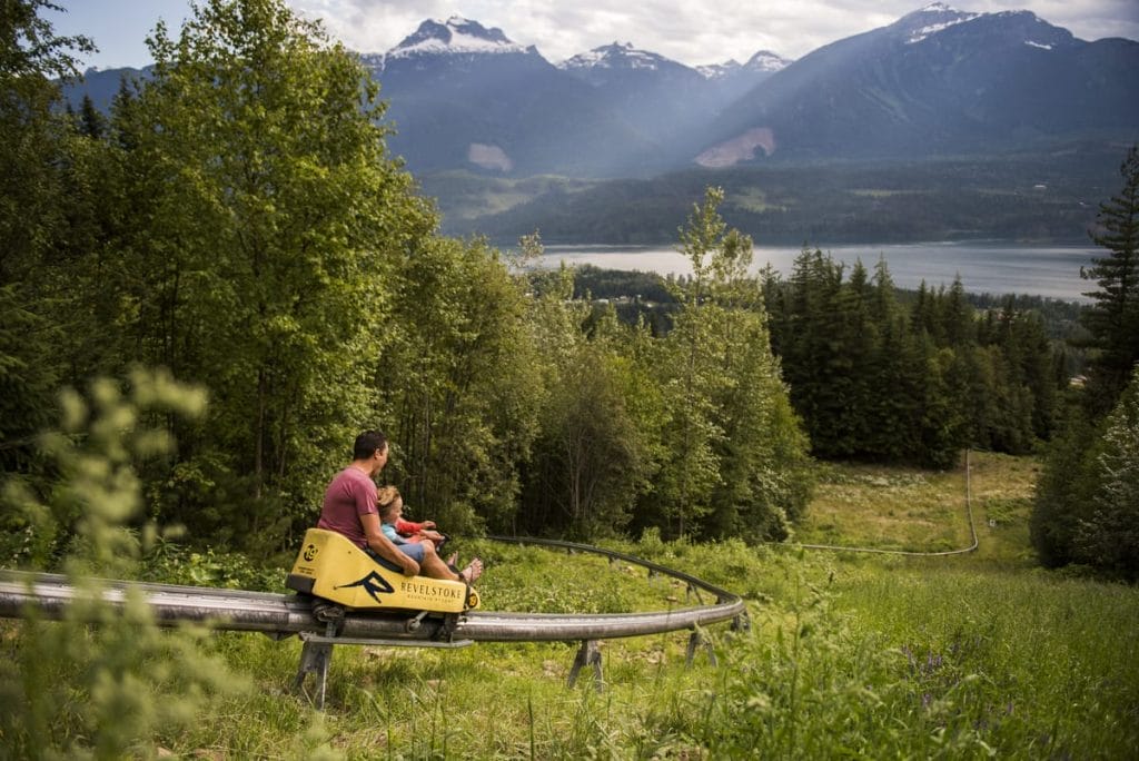 An adult and a child ride an alpine coaster down a green hillside, with mountains, forest, and a lake in the background—a perfect adventure during a staycation in BC.