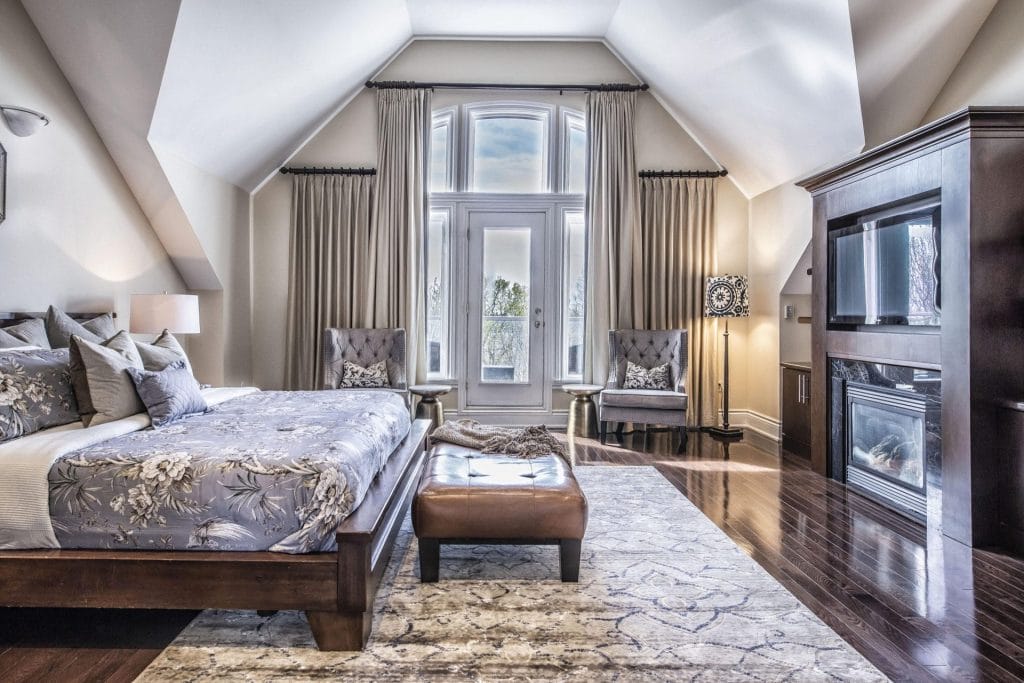 Spacious bedroom with vaulted ceiling, large bed, patterned rug, two chairs by tall windows with curtains—perfect to set up an Airbnb listing—plus a wall unit with TV and fireplace.