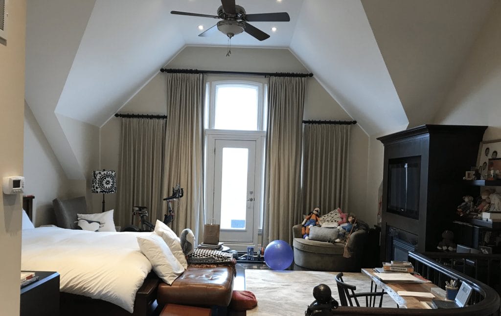 Spacious bedroom with vaulted ceiling—perfect to set up an Airbnb listing—features a bed on the left, central seating area with toys and exercise equipment, a TV unit on the right, and large windows with curtains at the back.