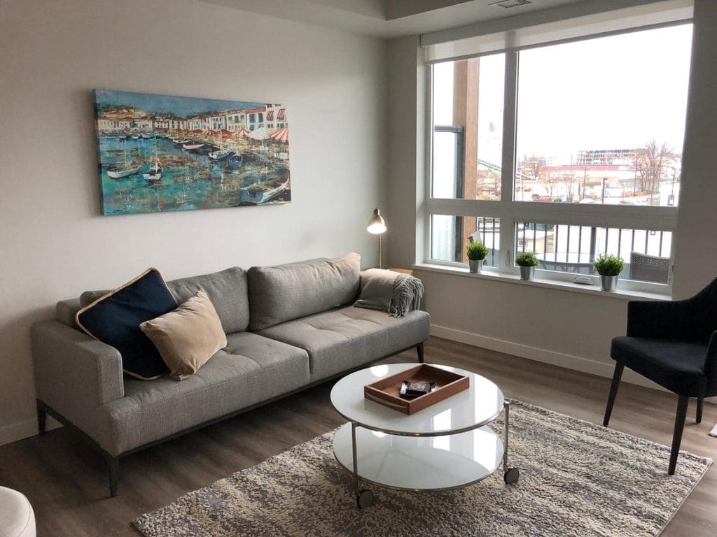 A modern living room with a gray sofa, two cushions, a small lamp, a round glass coffee table, and a painting above the sofa reflects the state of the industry in stylish home design, with a window displaying potted plants on the sill.