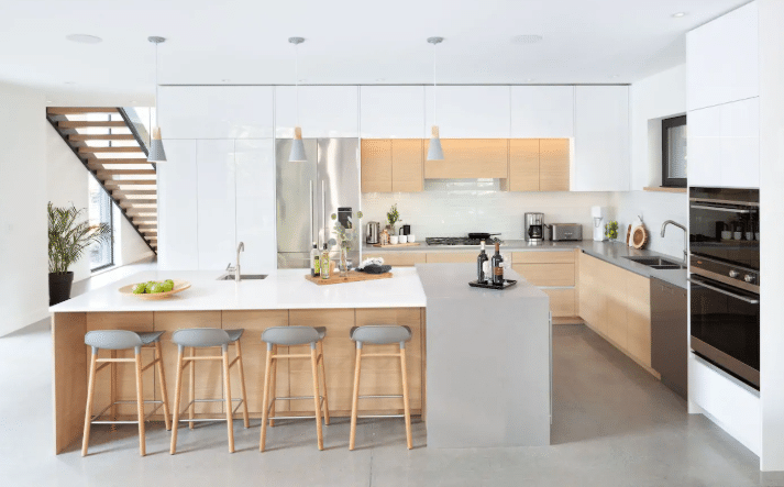 Modern kitchen with white and wood cabinets, island with four stools, stainless steel appliances, and pendant lights. A staircase is visible on the left side—showcasing a state-of-the-industry design.