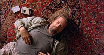 A man with long hair and a beard lies on his back on an ornate patterned rug, do it yourself Airbnb style, with one hand on his stomach and a notepad and cassette recorder nearby.