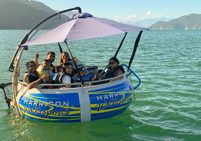 Six people sit and smile on a circular floating watercraft labeled "Harrison Watersports," with a canopy overhead, enjoying one of the top Things to Do in Harrison Hot Springs on a calm green lake surrounded by mountains.