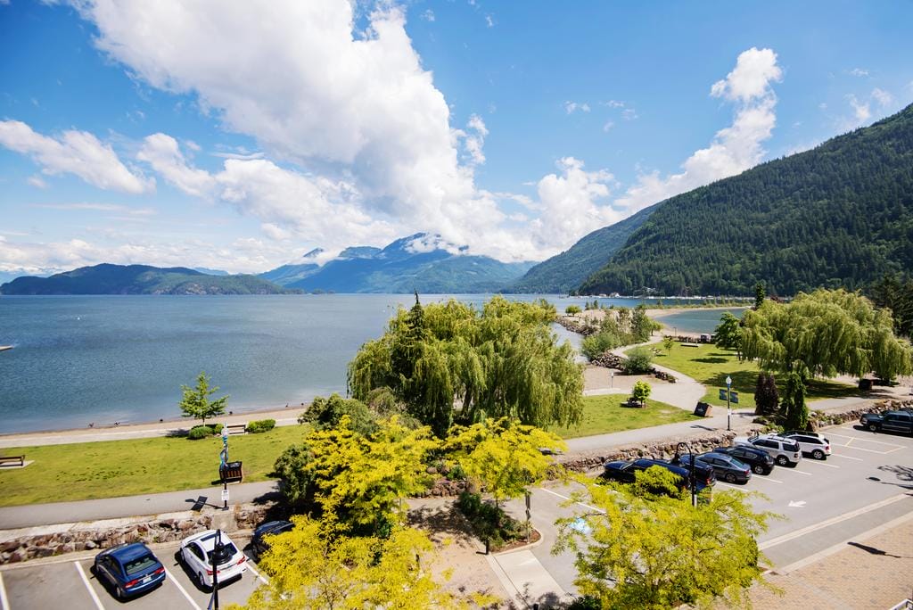 Enjoy a scenic lakeside view at Harrison Hot Springs, featuring mountains, a sandy beach, trees, parked cars, and a partly cloudy sky—perfect for relaxing and exploring the many things to do in Harrison Hot Springs.