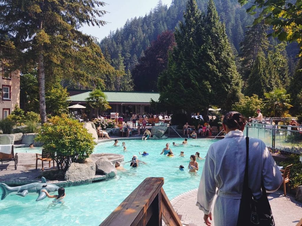 People relax and swim in an outdoor pool surrounded by trees and mountains, with a person in a robe standing in the foreground—one of the top things to do in Harrison Hot Springs.