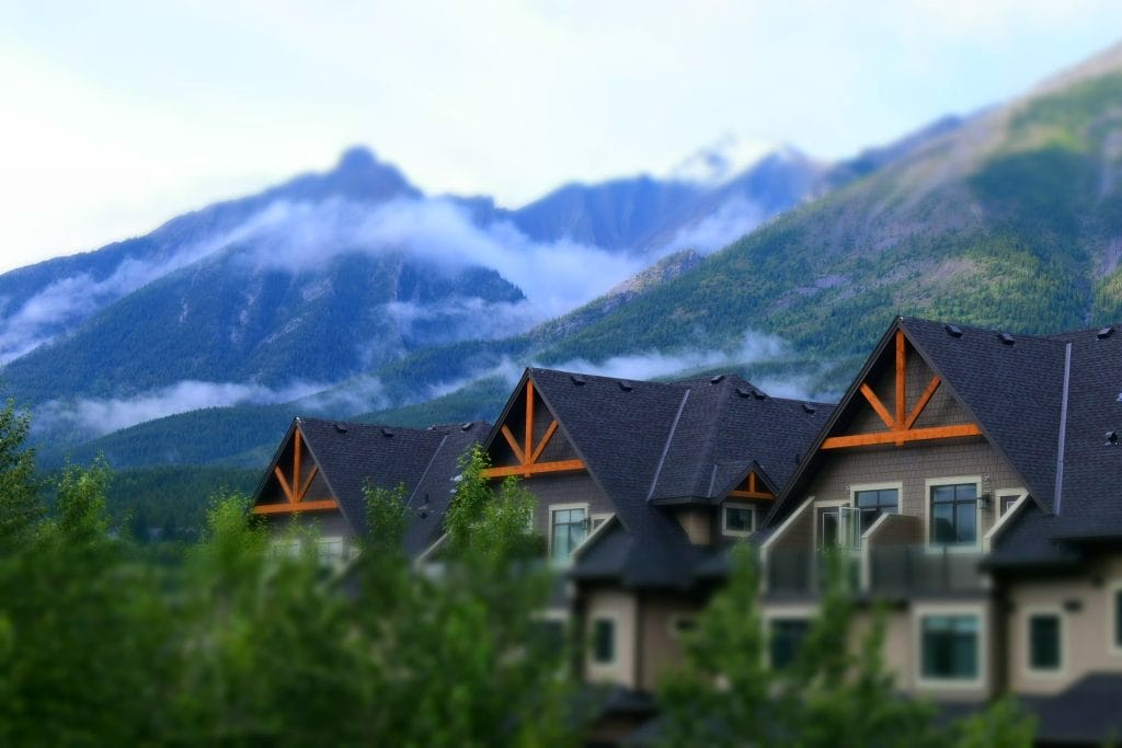 Several modern homes with dark roofs, popular in Airbnb markets in Canada, are set against a backdrop of green mountains with low-hanging clouds.