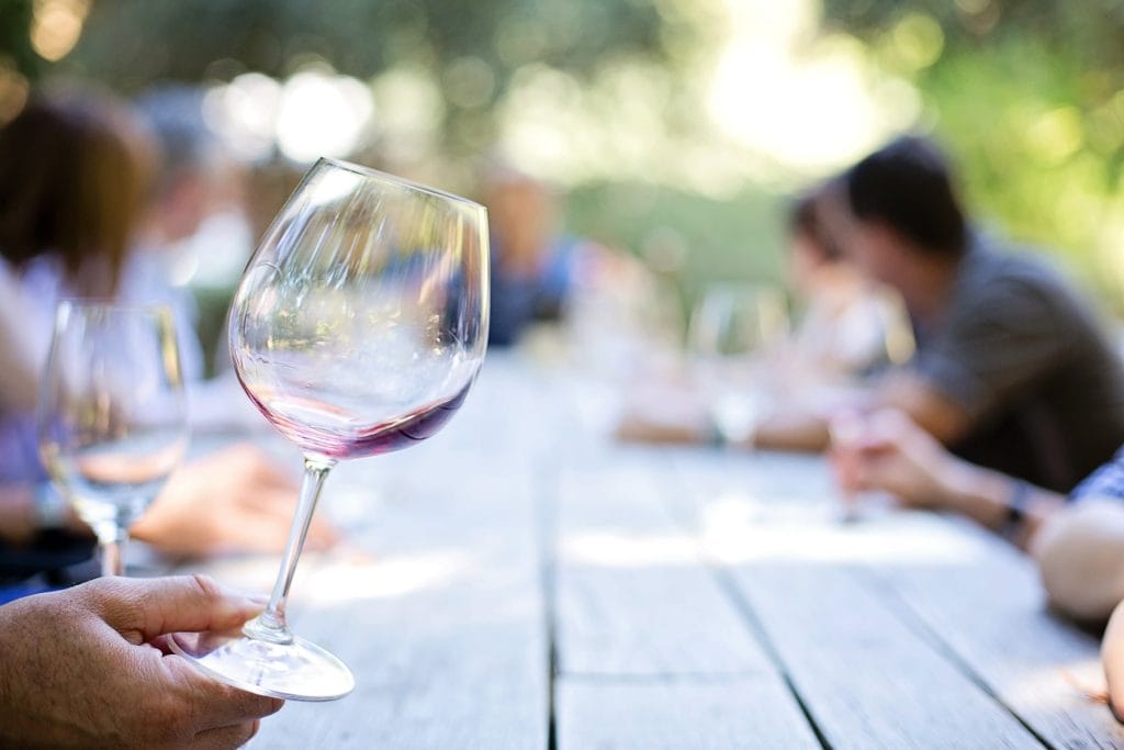 A hand holds a partially filled wine glass over an outdoor wooden table, with several people sitting and talking in the blurred background—evoking the relaxed charm found in popular Airbnb markets in Canada.