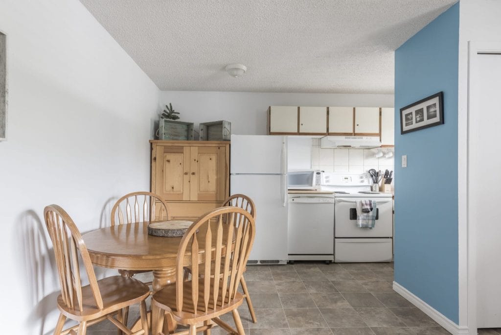 A small kitchen with white appliances, light wood cabinets, a round dining table with four chairs, and a blue accent wall—perfectly styled for Airbnb photography.