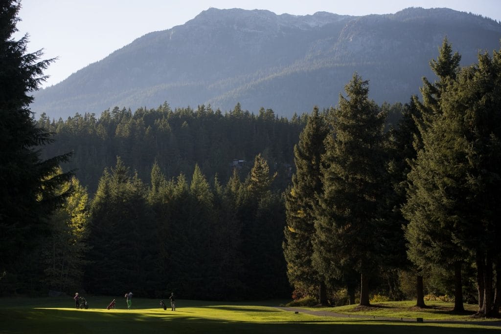 A group of people play golf on a green surrounded by tall pine trees, with a mountain range in the background under a clear sky, capturing the scenic beauty of golf courses in Whistler.