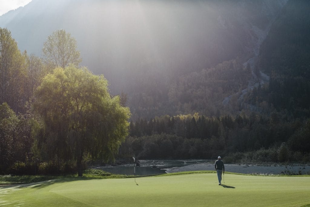 A person stands on a golf course green near a flag, surrounded by trees and mountains under soft sunlight, capturing the beauty found at golf courses in Whistler.