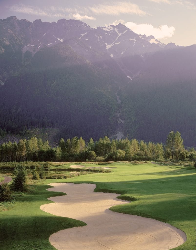 A golf course in Whistler with sand bunkers in the foreground, green fairways, and tall forested mountains in the background under soft sunlight.