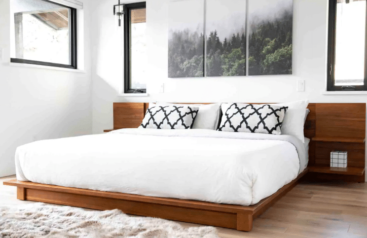 A modern bedroom perfect for Airbnb photography, featuring a wooden platform bed, crisp white bedding, two patterned pillows, wall art above the bed, and windows on both sides.