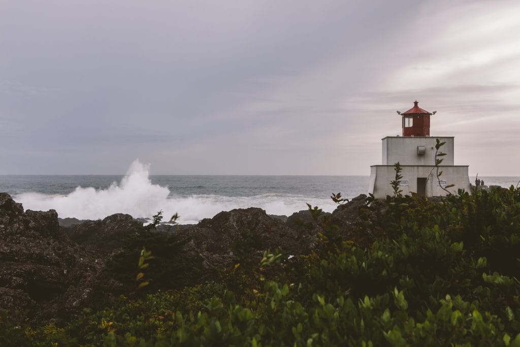 A white lighthouse with a red top stands on the rocky coastline near Ucluelet as ocean waves crash nearby under a cloudy sky. Shrubs and greenery are visible in the foreground.