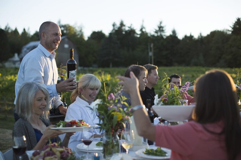 A group of people sit at a long outdoor table with food and drinks as a man pours wine; flowers and greenery, typical of Airbnbs on BC farms, are visible in the background.