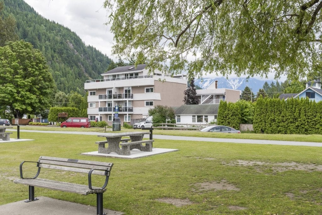 A park with a wooden bench and concrete picnic tables sits in the foreground, while an apartment building—ideal for Vacation Rental Management in Harrison Hot Springs—and mountains rise in the background.