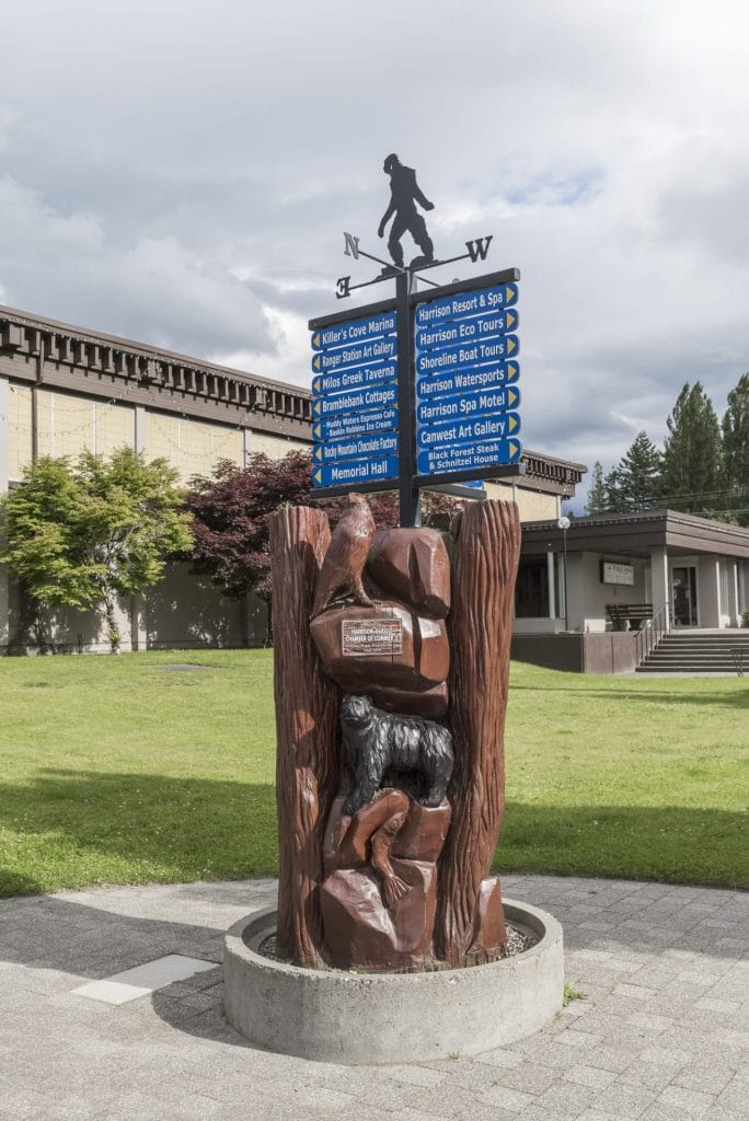 A wooden Sasquatch statue holds tourist direction signs in front of a building, with green grass and cloudy sky in the background—a quirky sight while exploring Vacation Rental Management in Harrison Hot Springs.