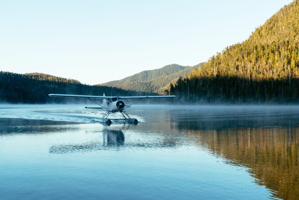 A small seaplane lands on a calm lake surrounded by forested hills and mountains under a clear sky, offering a glimpse of the scenic adventures among the top things to do in Ucluelet.